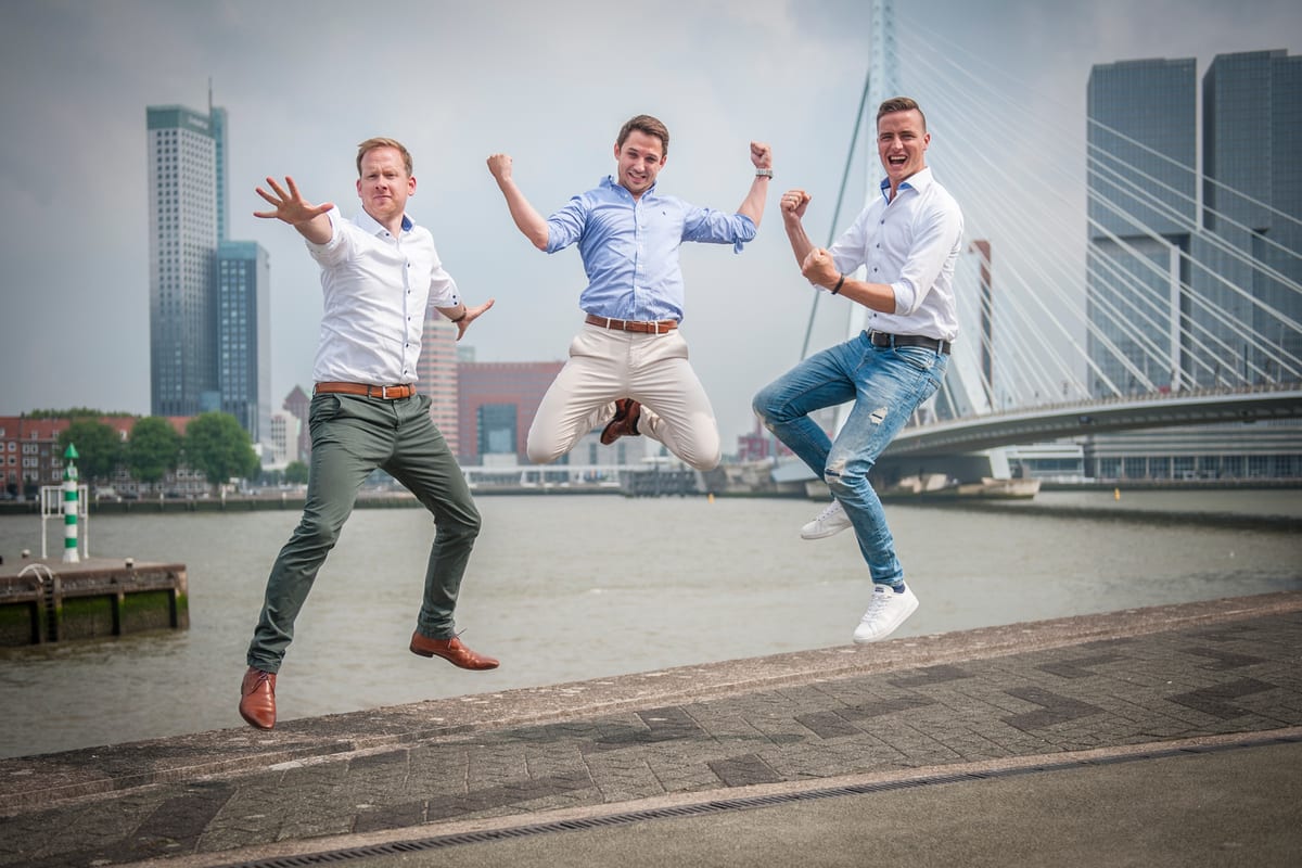 Drie mannen in wit hemd springen tegelijk omhoog op een kade, met de Erasmusbrug en de Rotterdamse skyline achter zich